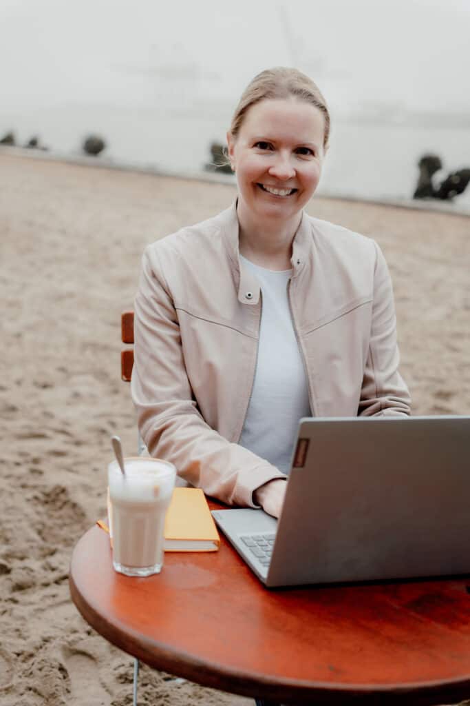 Content-Marketing-Beratung - Simone Maader am Elbstrand, am Tisch sitzend und lächelnd, mit Laptop, Kaffeebecher und gelbem Notizbuch